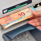 Eine Hand hebt Geldscheine, darunter ein 10-Euro-Schein, aus einem Geldautomaten ab. Der grüne Balken über "GELD" zeigt den erfolgreichen Vorgang an.