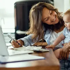 Lächelnde Mutter mit Baby auf dem Arm schreibt am Laptop. Sie plant die Zukunft und legt für das kinder sparbuch.