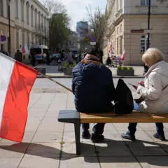 Zwei Personen sitzen auf einer Bank, eine polnische Flagge weht im Wind. Im Hintergrund sind Gebäude und Menschen zu sehen, die an einer Demonstration teilnehmen. Vielleicht geht es um den **ETF Polen**.