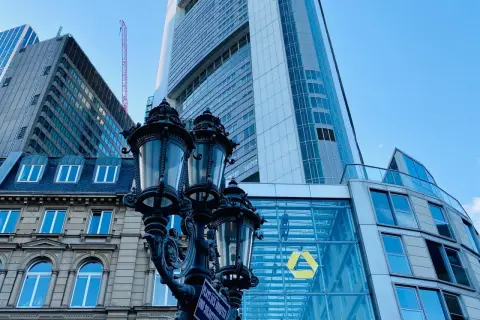 Ein historischer Laternenpfahl vor dem modernen Commerzbank-Gebäude in Frankfurt. Die Fassade spiegelt den blauen Himmel wider.