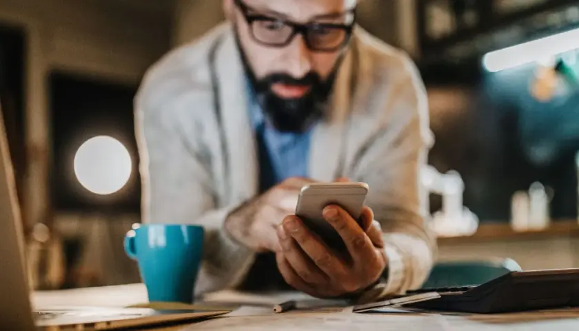Mann mit Brille und Bart tippt auf Smartphone. Neben ihm eine blaue Tasse und ein Taschenrechner. Er sucht vielleicht, was ist die bankleitzahl.