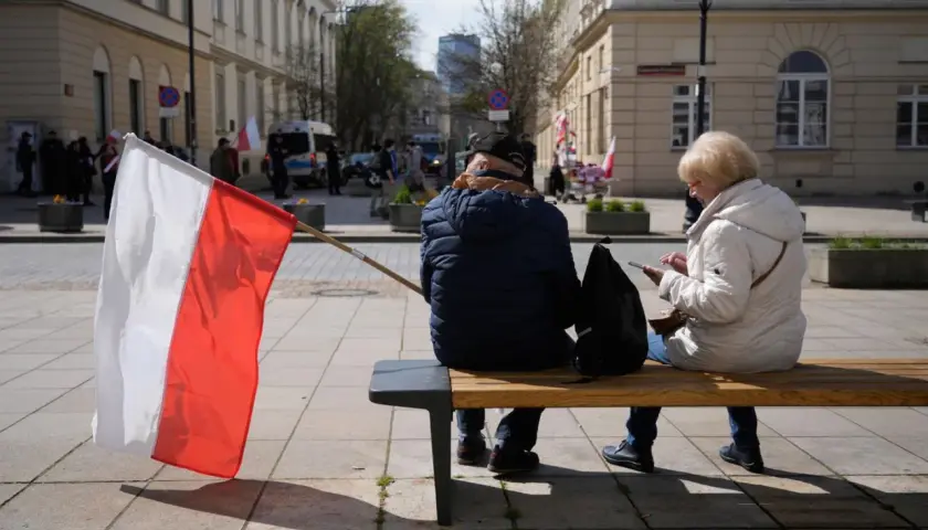 Zwei Personen sitzen auf einer Bank, eine polnische Flagge weht im Wind. Im Hintergrund sind Gebäude und Menschen zu sehen, die an einer Demonstration teilnehmen. Vielleicht geht es um den **ETF Polen**.