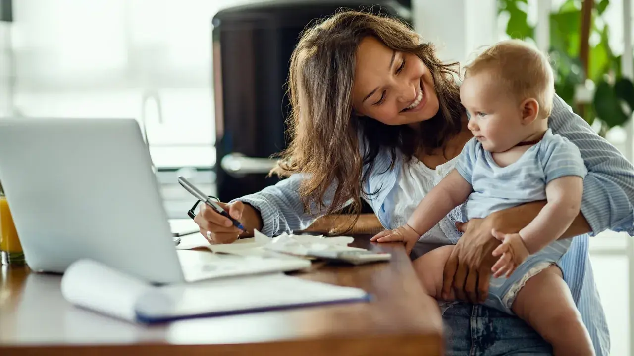 Lächelnde Mutter mit Baby auf dem Arm schreibt am Laptop. Sie plant die Zukunft und legt für das kinder sparbuch.