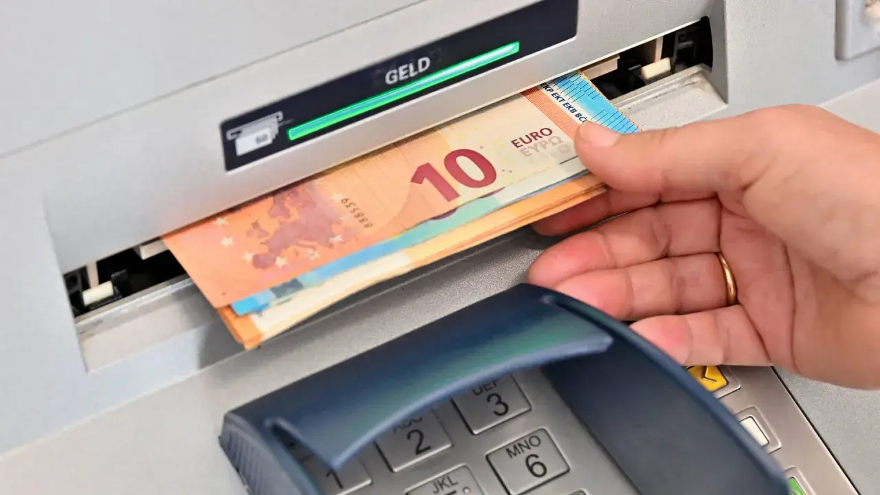 Eine Hand hebt Geldscheine, darunter ein 10-Euro-Schein, aus einem Geldautomaten ab. Der grüne Balken über "GELD" zeigt den erfolgreichen Vorgang an.