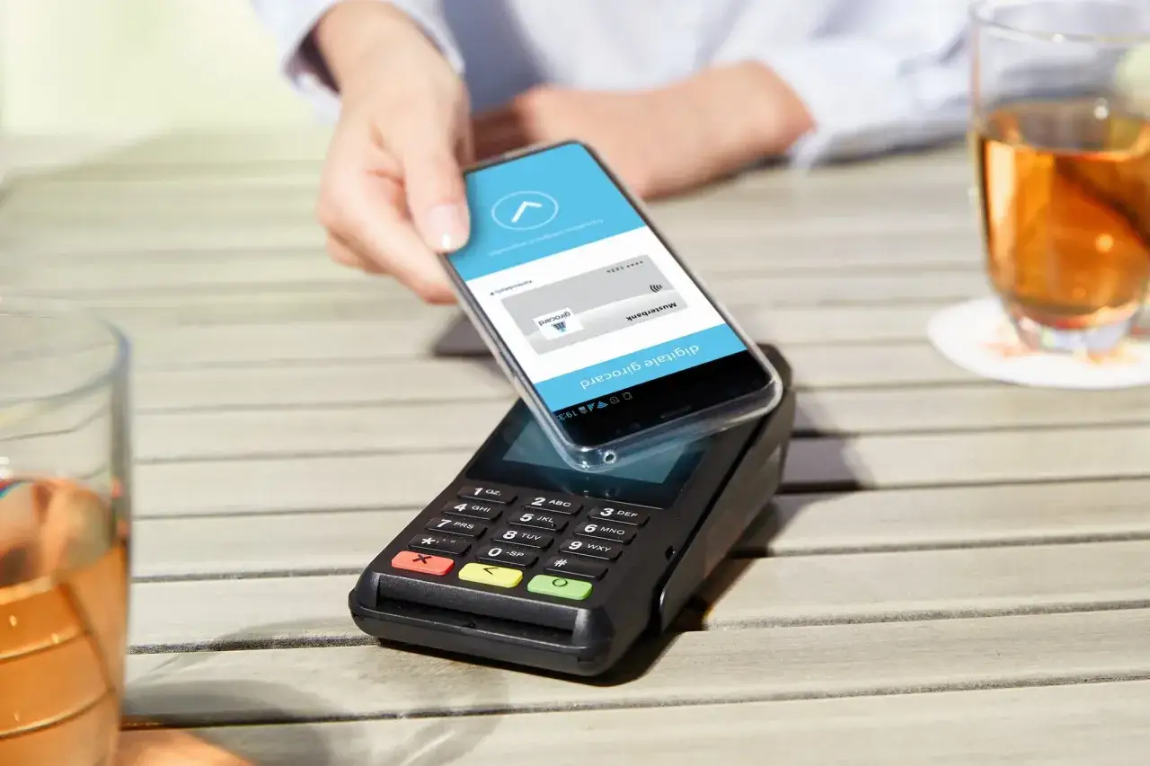 Eine Hand h&auml;lt ein Smartphone &uuml;ber ein Kassenterminal, um mit einer digitalen girocard zu bezahlen.